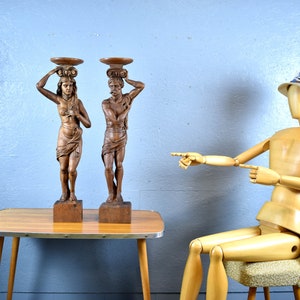 Puede incluir: Dos esculturas de madera de personas que llevan cestas en la cabeza. Las esculturas son de color marr&oacute;n y tienen un acabado r&uacute;stico. Est&aacute;n de pie sobre una mesa de madera con un acabado de madera clara. Un maniqu&iacute; de madera est&aacute; sentado en una silla en el fondo, se&ntilde;alando las esculturas.