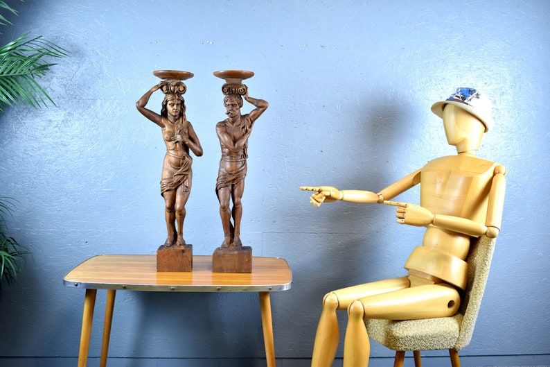 Puede incluir: Dos esculturas de madera de personas que llevan cestas en la cabeza. Las esculturas son de color marr&oacute;n y tienen un acabado r&uacute;stico. Est&aacute;n de pie sobre una mesa de madera con un acabado de madera clara. Un maniqu&iacute; de madera est&aacute; sentado en una silla en el fondo, se&ntilde;alando las esculturas.