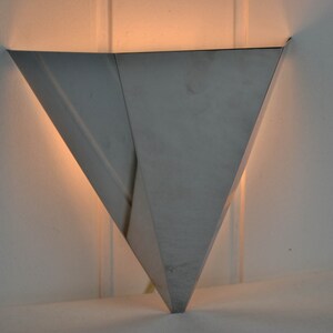 May include: A silver triangular wall sconce with a light shining from inside.