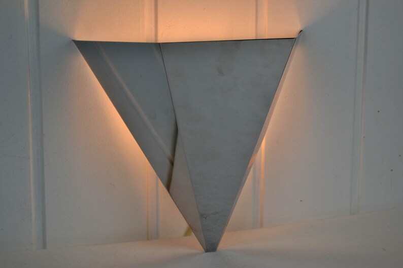May include: A silver triangular wall sconce with a light shining from inside.