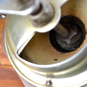 K&ouml;nnte beinhalten: Nahaufnahme des Inneren einer Vintage-Kaffeem&uuml;hle. Die M&uuml;hle ist aus Metall und hat einen Holzsockel. Der Mahlmechanismus ist sichtbar und es befinden sich Kaffeereste in der M&uuml;hle.