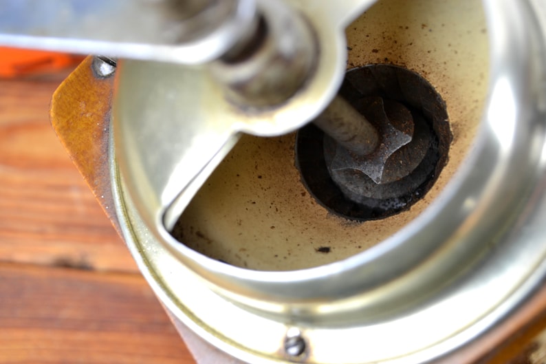 K&ouml;nnte beinhalten: Nahaufnahme des Inneren einer Vintage-Kaffeem&uuml;hle. Die M&uuml;hle ist aus Metall und hat einen Holzsockel. Der Mahlmechanismus ist sichtbar und es befinden sich Kaffeereste in der M&uuml;hle.