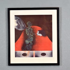 May include: A framed print of a surrealist painting featuring a green, humanoid figure with a large, red, abstract shape behind it. A woman's face is partially visible in the red shape.