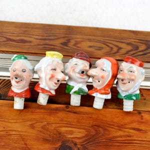 May include: Six ceramic bottle stoppers with whimsical faces. Each stopper features a different character with unique facial expressions and colorful clothing. The stoppers are arranged in a row on a wooden surface.