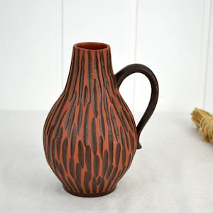 May include: A brown ceramic vase with a textured, striped design and a handle. The vase has a rounded body and a narrow neck.