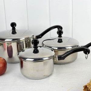 May include: A set of three stainless steel saucepans with black handles and lids. The saucepans are of different sizes and are arranged on a white surface.