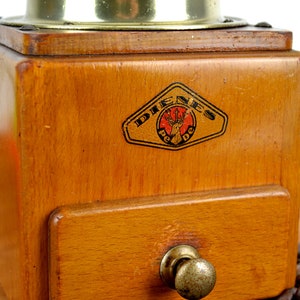 K&ouml;nnte beinhalten: Antiker Kaffeem&uuml;hle aus Holz mit Messingdeckel und Schublade. Die M&uuml;hle tr&auml;gt ein Etikett mit der Aufschrift "Dienes Pe De" und einem Hirschlogo.