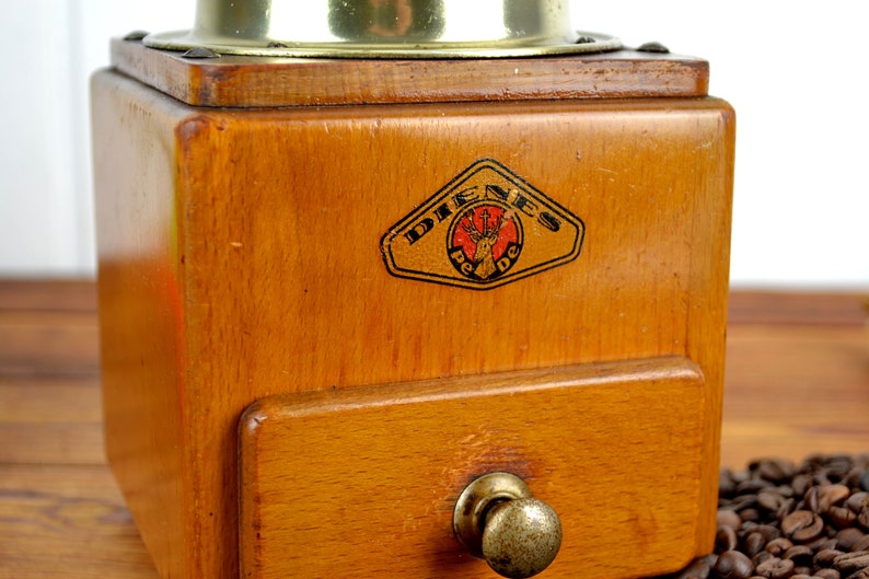 K&ouml;nnte beinhalten: Antiker Kaffeem&uuml;hle aus Holz mit Messingdeckel und Schublade. Die M&uuml;hle tr&auml;gt ein Etikett mit der Aufschrift "Dienes Pe De" und einem Hirschlogo.