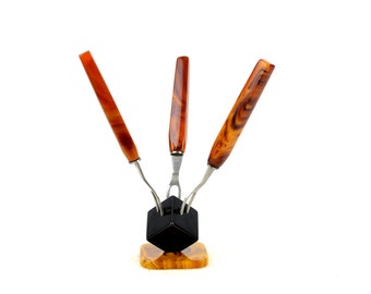 May include: A set of three fondue forks with stainless steel tines and marbled orange and brown handles. The forks are displayed in a black and orange stand with a square base, against a white background.