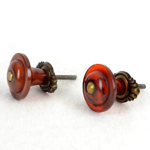 Peut inclure: Deux boutons de placard ronds présentant une surface en résine polie de couleur ambre marbré avec un centre lisse et bombé. Chaque bouton comprend une petite incrustation métallique circulaire couleur laiton au centre et une base en laiton antique cannelée.