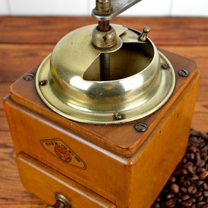 K&ouml;nnte beinhalten: Eine antike Kaffeem&uuml;hle aus Holz mit Messingdeckel und Kurbel. Die M&uuml;hle hat eine Schublade zum Auffangen des Kaffeesatzes und ein Etikett mit der Aufschrift "Die  " auf Deutsch. Die M&uuml;hle steht auf einem Haufen Kaffeebohnen.