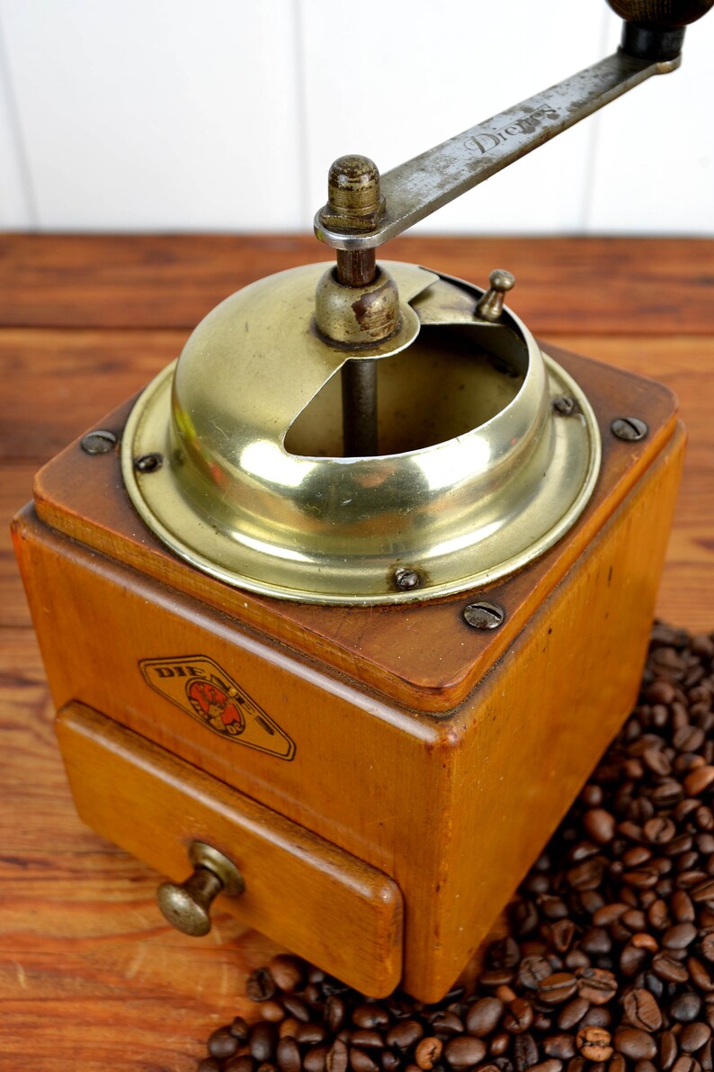 K&ouml;nnte beinhalten: Eine antike Kaffeem&uuml;hle aus Holz mit Messingdeckel und Kurbel. Die M&uuml;hle hat eine Schublade zum Auffangen des Kaffeesatzes und ein Etikett mit der Aufschrift "Die  " auf Deutsch. Die M&uuml;hle steht auf einem Haufen Kaffeebohnen.