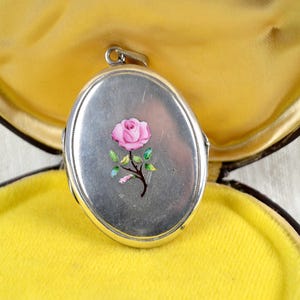 May include: Silver oval locket with a pink rose design, set in a yellow velvet-lined box. The locket features a detailed floral illustration. The box is dark brown with a gold-colored trim.