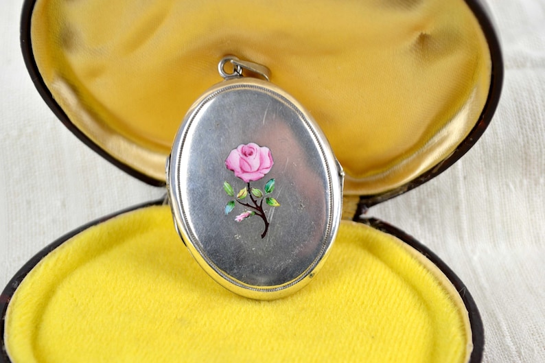 May include: Silver oval locket with a pink rose design, set in a yellow velvet-lined box. The locket features a detailed floral illustration. The box is dark brown with a gold-colored trim.