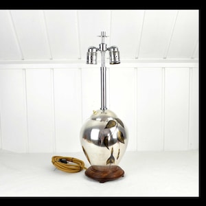 May include: A silver-toned table lamp with a bulbous base and a chrome stem. The base features a painted leaf design. The lamp sits on a round, dark wooden base. A brown electrical cord is coiled on the surface.
