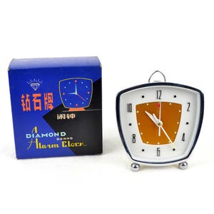 May include: Vintage Diamond Alarm Clock with a white face, orange center, and black trim. The clock has silver feet and a loop for hanging. A blue box with Chinese characters and the words "Diamond Alarm Clock" is next to the clock.