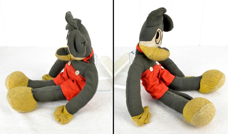 May include: A vintage plush Mickey Mouse toy with black fur, red shorts, and yellow shoes. The toy is sitting with its legs outstretched and its head turned to the side.