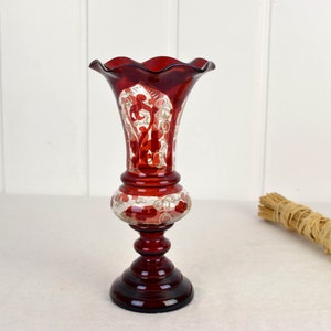 May include: A red glass vase with a white floral design. The vase has a scalloped rim and a flared base.