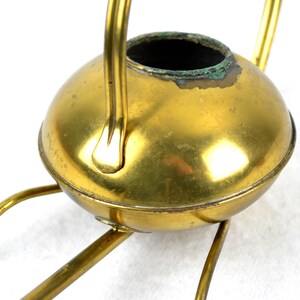 May include: A gold-toned metal watering can with a round body and a long spout. The can has a handle and sits on a three-legged stand.