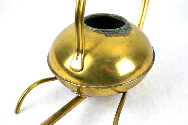 May include: A gold-toned metal watering can with a round body and a long spout. The can has a handle and sits on a three-legged stand.