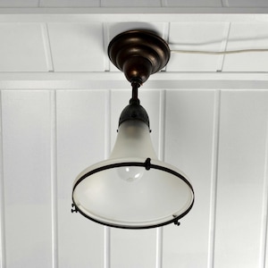 Op de afbeelding: Een witte, gematteerde glazen plafondlamp met een zwart metalen frame en een zwart metalen kap. De lamp hangt aan een wit plafond.