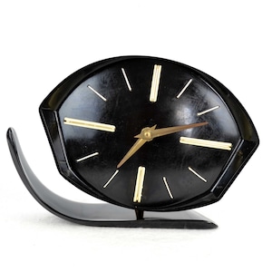 May include: A black, mid-century modern clock with a unique, curved base. The clock face is black with gold-colored hour markers and hands.