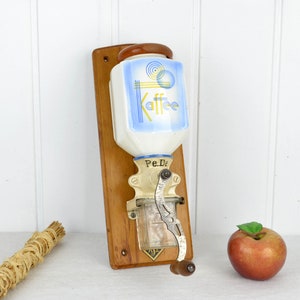 Può includere: Macinacaffè vintage da parete con un barattolo in ceramica blu e bianco che dice "Kaffee" e una cornice in legno. Il macinacaffè ha una manovella in metallo e un barattolo di vetro per il caffè macinato.