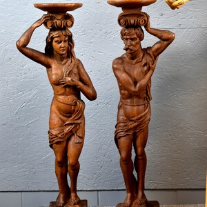 Puede incluir: Un par de esculturas de madera de un hombre y una mujer, cada uno sosteniendo una bandeja en la cabeza. Las esculturas est&aacute;n talladas en un estilo realista y est&aacute;n pintadas en un acabado de madera natural.