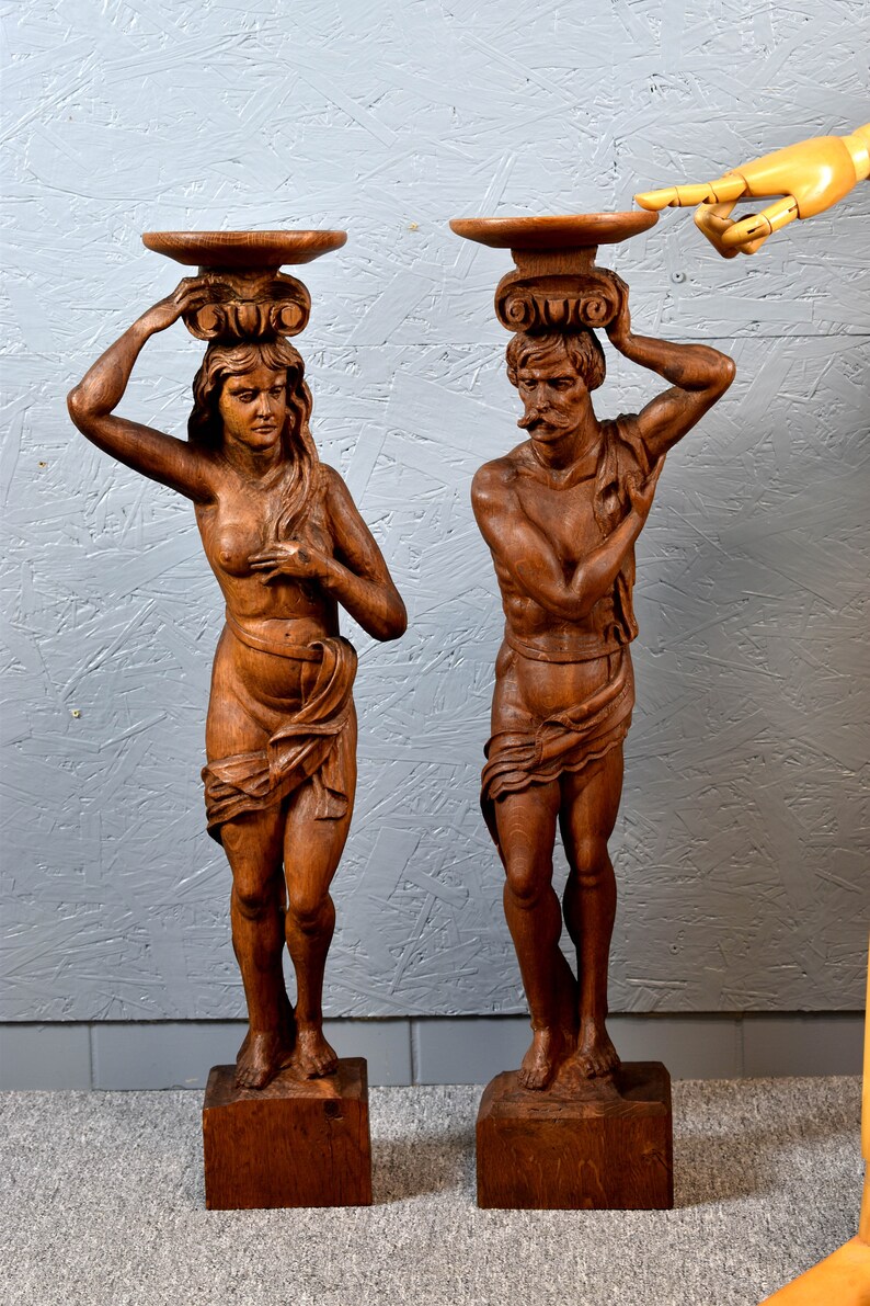 Puede incluir: Un par de esculturas de madera de un hombre y una mujer, cada uno sosteniendo una bandeja en la cabeza. Las esculturas est&aacute;n talladas en un estilo realista y est&aacute;n pintadas en un acabado de madera natural.