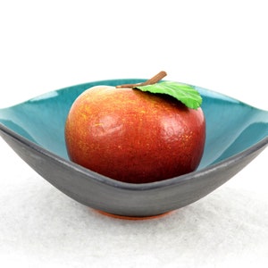 May include: A red apple with a green leaf sits in a teal and black ceramic bowl. The bowl has a square shape with rounded edges.