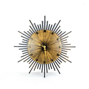 May include: A gold and black sunburst clock with a woodgrain face and black hour and minute hands.
