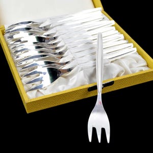 Könnte beinhalten: Ein Set silberner Gabeln in einer gelben Schachtel mit weißem Satinfutter. Eine Gabel wird vor der Schachtel präsentiert. Die Gabeln haben ein dreizinkiges Design und eine elegante, moderne Ästhetik, perfekt für formelle Anlässe.
