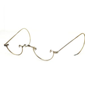 May include: Antique, wire-rimmed eyeglasses with a delicate, curved design. The glasses feature a bridge and curved temples, and are made of a light-colored metal. The lenses are missing, and the glasses are set against a white background.