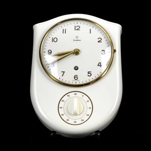 Könnte beinhalten: Eine weiße Wanduhr mit glänzender Oberfläche, bestehend aus einem runden Zifferblatt mit schwarzen Ziffern und goldfarbenen Zeigern. Unter dem Zifferblatt befindet sich ein runder Timer mit weißem Drehknopf, markiert von 0 bis 60. Das Junghans-Logo ist sichtbar.