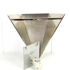 May include: A silver metal wall sconce with a triangular shape and a flat back plate. The sconce has a simple design and is made of polished metal.