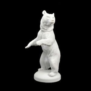 May include: A white ceramic bear figurine standing upright on a circular base. The bear has a detailed texture and is posed with one arm raised. The sculpture is set against a black background, highlighting its form.