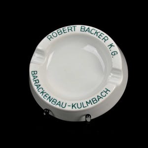 Könnte beinhalten: Weißer Keramik-Aschenbecher mit dem Text "ROBERT BACKER K.G." und "BARACKENBAU-KULMBACH" in grüner Schrift. Der Aschenbecher hat ein kreisförmiges Design mit einer vertieften Mitte und drei Kerben.