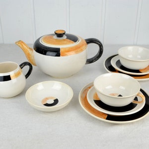 Op de afbeelding: Een vintage theeset met een crèmekleurige basis en geometrische zwarte, oranje en bruine accenten. De set bevat een theepot, een melkkan, een suikerpot en twee theekopjes met schotels.