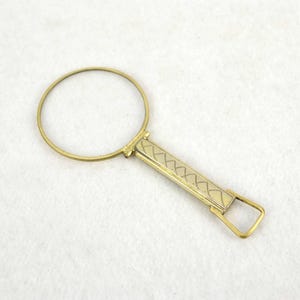 May include: A vintage brass magnifying glass with a circular lens and a rectangular handle. The handle features a decorative geometric pattern. The magnifying glass is designed for reading or examining small objects.