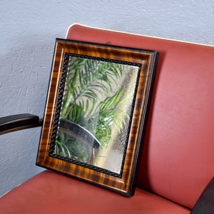 Könnte beinhalten: Ein rechteckiger Spiegel mit einem dunklen Holzmaserungsrahmen und einer schwarzen, seilartigen Zierleiste. Der Spiegel liegt auf einem roten Stuhl mit weißer Zierleiste. Der Hintergrund zeigt einen Blick auf grünes Laub.