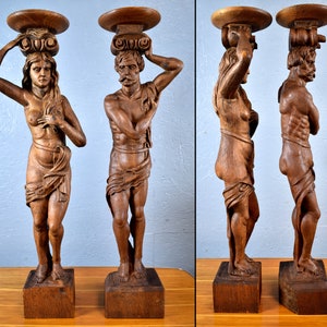 Puede incluir: Dos esculturas antiguas de madera de un hombre y una mujer, cada una de pie sobre un pedestal con una parte superior redonda. Las esculturas est&aacute;n talladas en un estilo realista y presentan detalles intrincados.