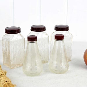 Op de afbeelding: Set van vijf heldere glazen flessen met kastanjebruine plastic deksels. De flessen hebben een getextureerd oppervlak en zijn gerangschikt op een wit oppervlak. Een rode appel is op de achtergrond.
