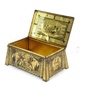 Puede incluir: Una caja de metal dorado con una tapa con bisagras y un diseño decorativo. La tapa presenta una escena de un carro tirado por caballos. La caja está abierta y el interior está vacío.