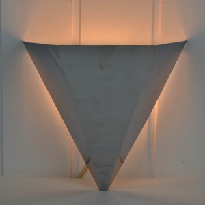 May include: A silver triangular wall sconce with a light shining through the opening.
