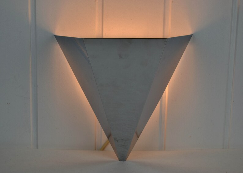 May include: A silver triangular wall sconce with a light shining through the opening.