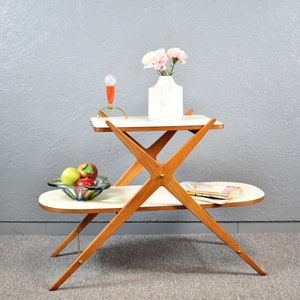May include: A two-tiered white table with a wooden frame. The table has a unique X-shaped design and is decorated with a vase of pink flowers and a bowl of fruit.