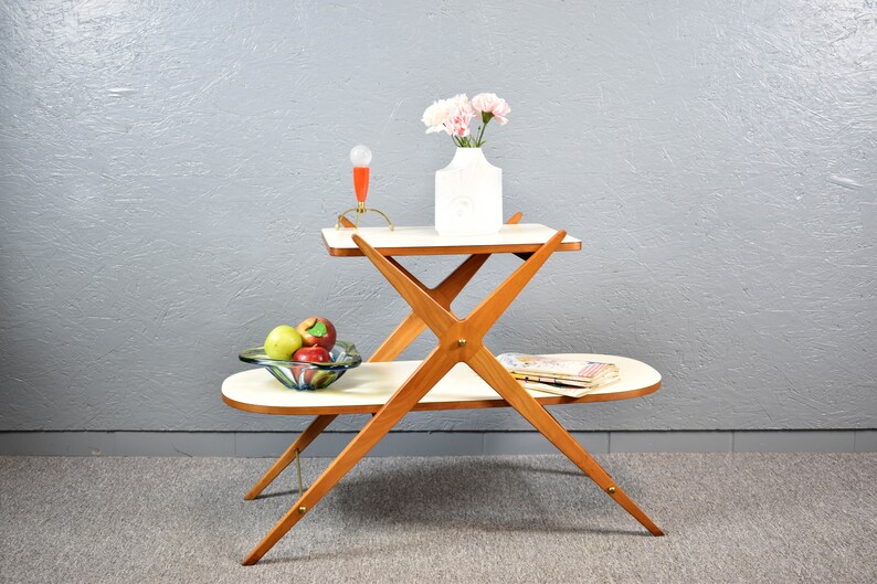 May include: A two-tiered white table with a wooden frame. The table has a unique X-shaped design and is decorated with a vase of pink flowers and a bowl of fruit.
