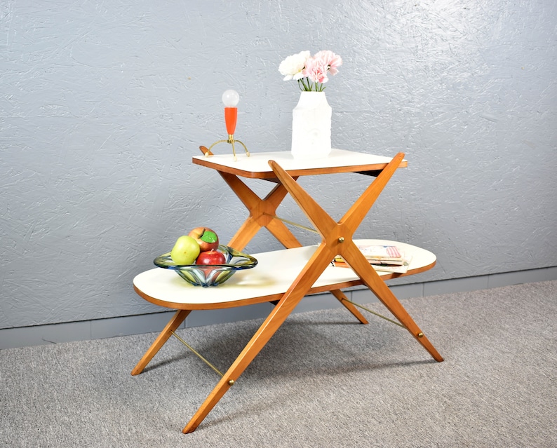 May include: A white two-tiered serving cart with a wooden frame. The cart has a white top shelf and a white oval-shaped bottom shelf. The cart is on a gray carpet. There is a glass bowl with fruit on the bottom shelf and a vase with pink flowers on the top shelf.
