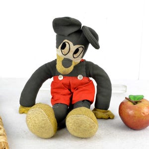 May include: Vintage plush Mickey Mouse toy with black fur, red trousers, and a black hat. The toy is sitting on a white surface with a brown stick and a red apple.
