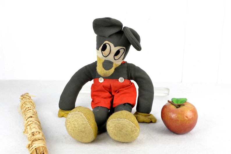 May include: Vintage plush Mickey Mouse toy with black fur, red trousers, and a black hat. The toy is sitting on a white surface with a brown stick and a red apple.